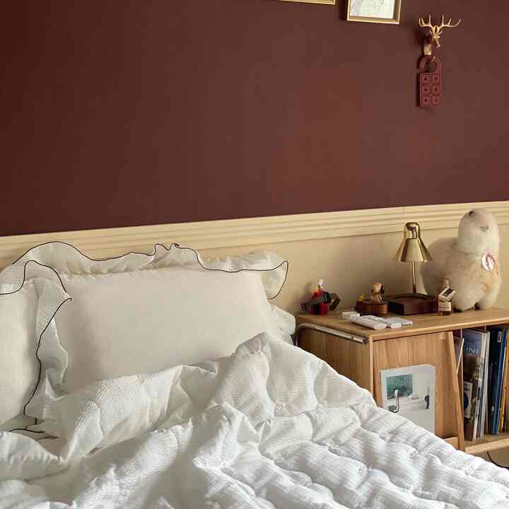 温かみのあるブラウンの壁と白い寝具が調和した寝室、木製のナイトテーブルとマガジンラックが配置された雰囲気のあるインテリア