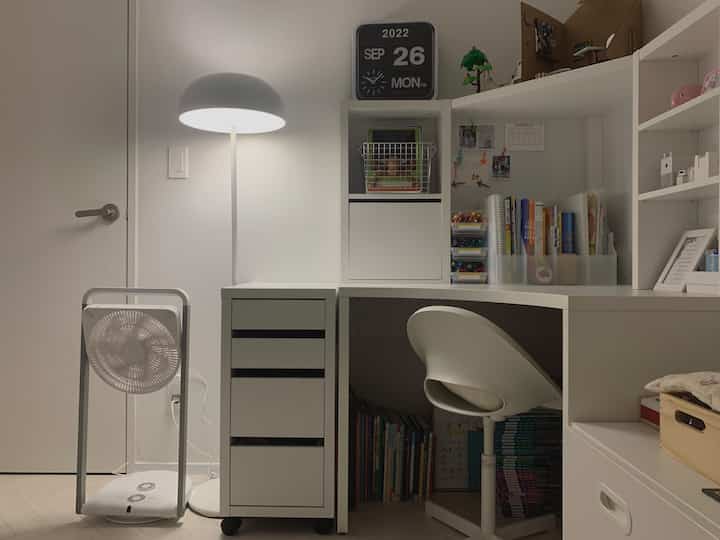 White-toned kids' room featuring a corner desk with chair, bookshelf, and floor lamp creating a neat study area