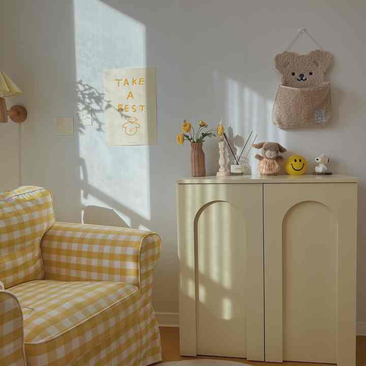 A cozy living room in yellow and beige tones, featuring a checkered sofa and a small storage cabinet with bear-themed details
