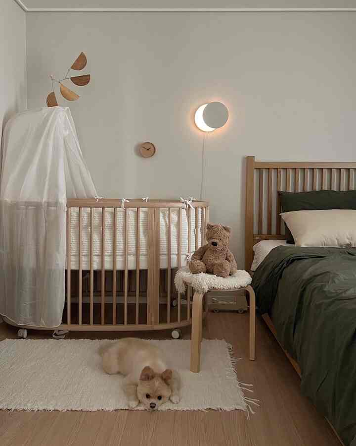Natural beige and khaki toned bedroom featuring a wooden crib, chair with teddy bear, and a pet dog lying on the rug