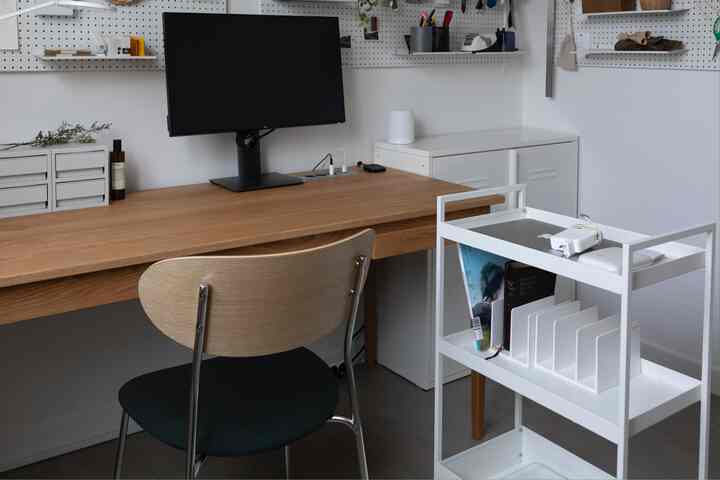 Clean home office with white walls and wood tone desk, featuring a monitor and a white storage trolley in a practical setup