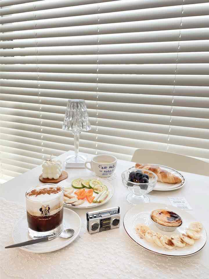 Bright white-toned dining room featuring blinds, a crystal table lamp, coffee drinks, and brunch plates creating a cozy home cafe atmosphere