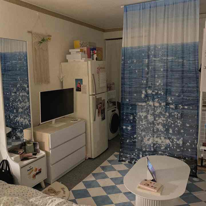 White and blue toned studio apartment living space featuring ocean-pattern curtain and simple furniture arrangement with cozy atmosphere