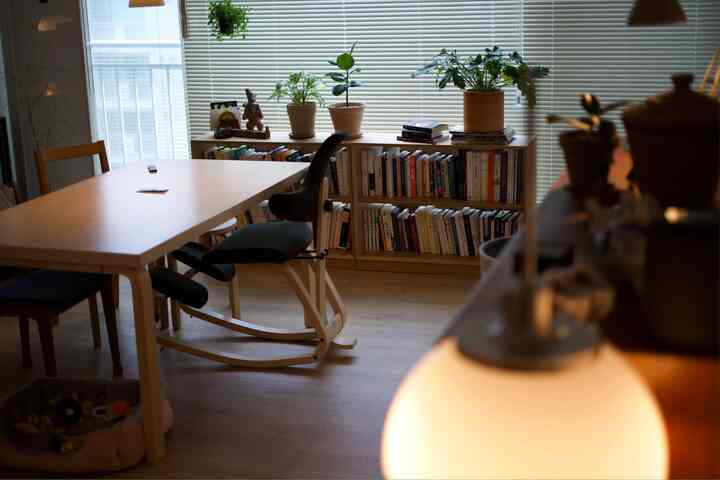 Living room with light wood tones and white blinds, featuring low bookshelf, dining table, and plants creating warm ambiance