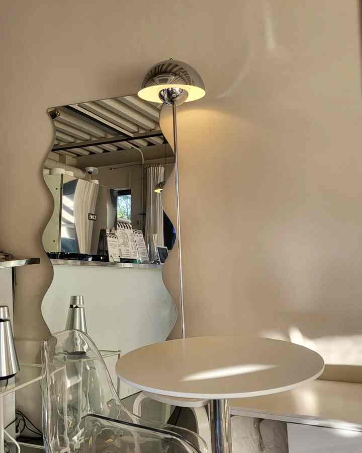 ベージュの壁に波型の鏡とシルバーのスタンドフロアランプ、白い丸テーブルが配置されたシンプルなカフェ風のインテリア
