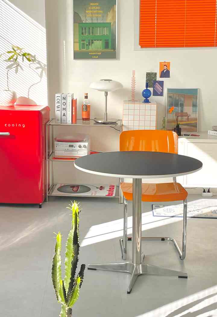 白い壁にオレンジのブラインド、レトロな赤いミニ冷蔵庫と丸い食卓、オレンジの椅子が配されたミッドセンチュリーモダンなキッチンスペース