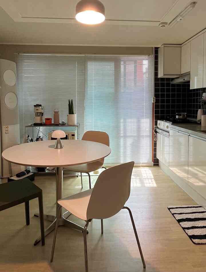 White and wood tone studio apartment kitchen and dining space featuring a round table and aluminum blinds in a clean, minimalist interior