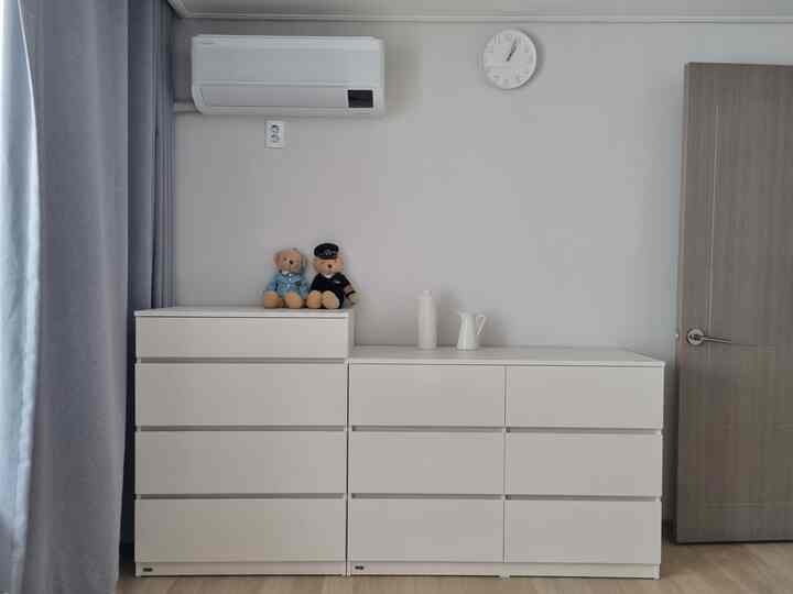 A calm bedroom in white and gray tones featuring two white dressers at the center, gray blackout curtains on the left, and a wall clock and air conditioner mounted above