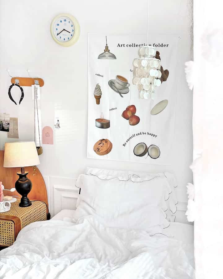 A small bedroom featuring white and brown tones, with a white bed with pillows in center, white curtain on right, and brown lamp and wicker bedside table on left creating cozy atmosphere