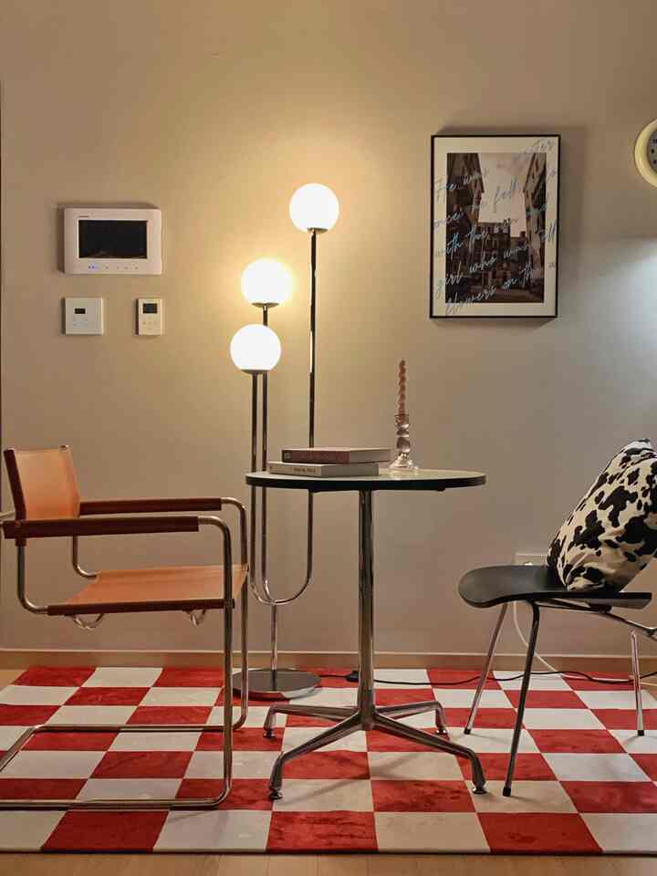 赤と白のチェッカー柄ラグが目を引くダイニング、ミッドセンチュリーモダンの円形テーブルと多様な椅子、暖かな照明が配置された雰囲気のあるインテリア