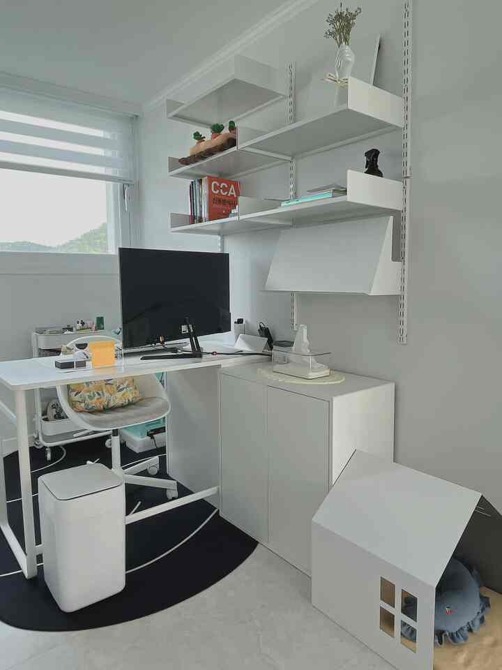 白を基調としたワンルームのホームオフィス、すっきりとしたデスクとチェア、壁の棚、犬小屋が配置されたモダンな雰囲気の空間