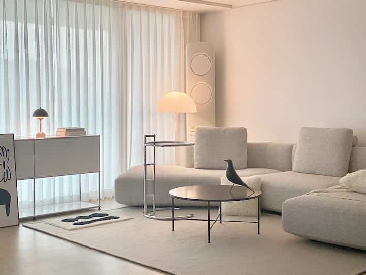 Natural tone living room featuring beige sectional sofa, sheer curtains, round coffee table, and floor lamp in a clean, minimalist setting