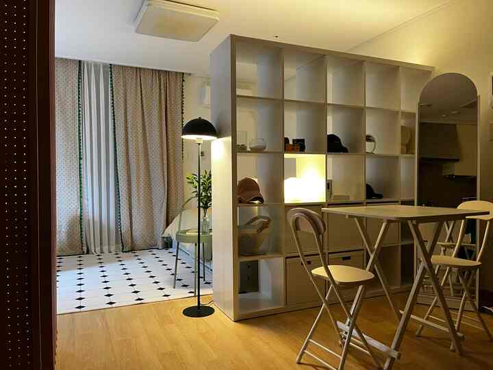 White and wood tone studio apartment of 9 pyeong featuring a cozy living space separated by a central KALLAX bookshelf room divider and folding table setup