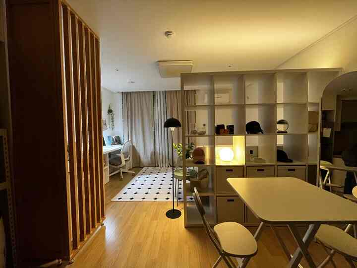 Warm wood tones and white dominate this kitchen area featuring a room divider bookshelf, folding dining table, and ambient lighting creating a cozy atmosphere