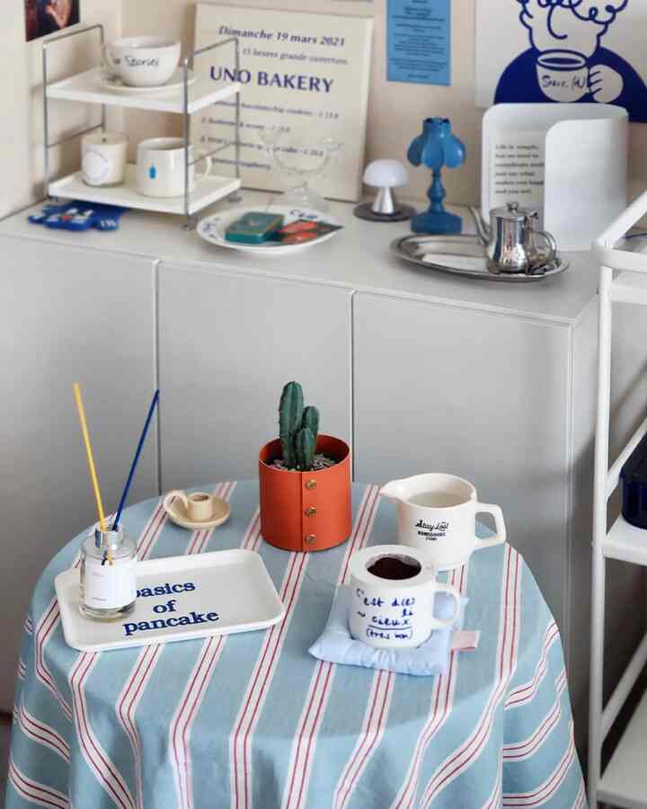 白い壁とキャビネットのあるキッチン、中央にブルーと赤のストライプのテーブルクロスをかけた丸テーブルとホームカフェの小物が配置されたシンプルでナチュラルな印象的な空間