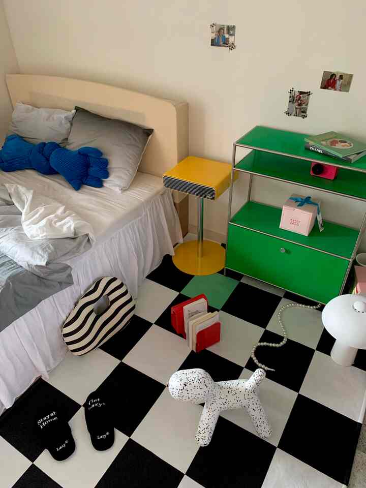 Modern bedroom with white walls and black and white checkerboard floor, featuring beige bed, yellow stool, and green storage unit