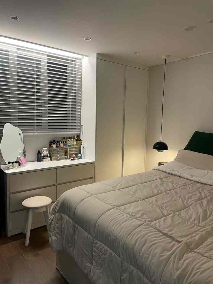 ホワイトと木目調のモダンな寝室、窓辺にチェストが置かれ、ブラインドが窓を覆う雰囲気のあるインテリア