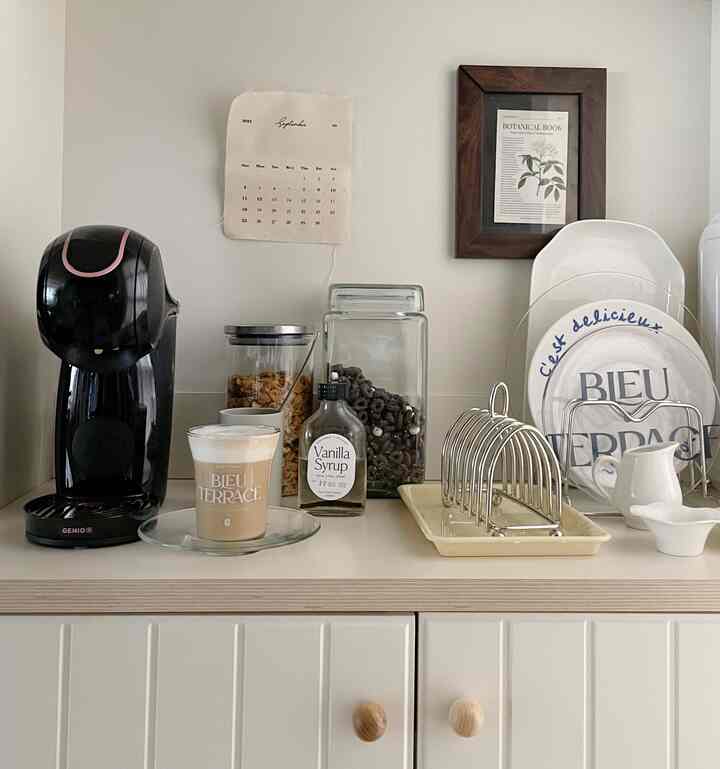 ベージュとホワイトの色調のキッチン空間、黒いカプセルコーヒーマシンと複数のコーヒー用品が配置された温かみのあるおうちカフェ