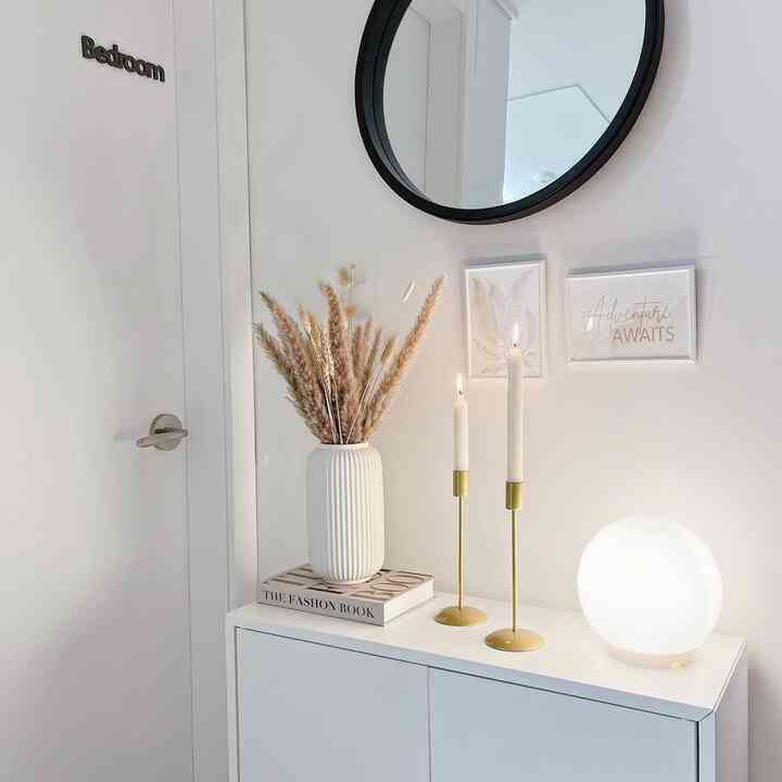 Bright white-toned bedroom entrance featuring a cabinet with natural brown pampas grass vase, gold candle holders, and a mood lamp creating a cozy atmosphere