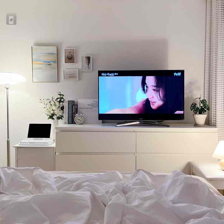 White-toned bedroom featuring a chest of drawers with TV, framed wall art, floor lamp, and a small desk creating a neat and cozy atmosphere