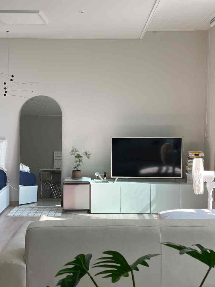 A white and natural-toned studio apartment featuring a full-length mirror, sofa, and storage cabinet in a clean living and bedroom area