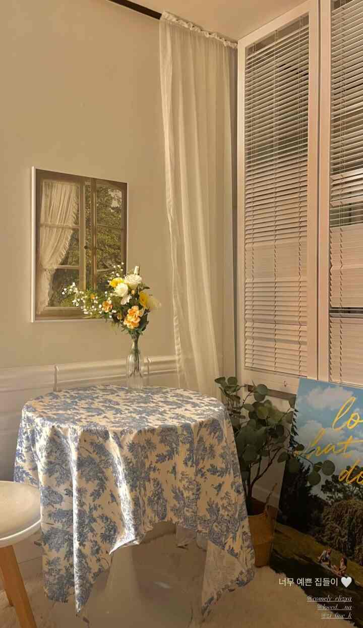 Beige-toned walls with white curtains, floral tablecloth on round table, and a plant in a cozy dining room space