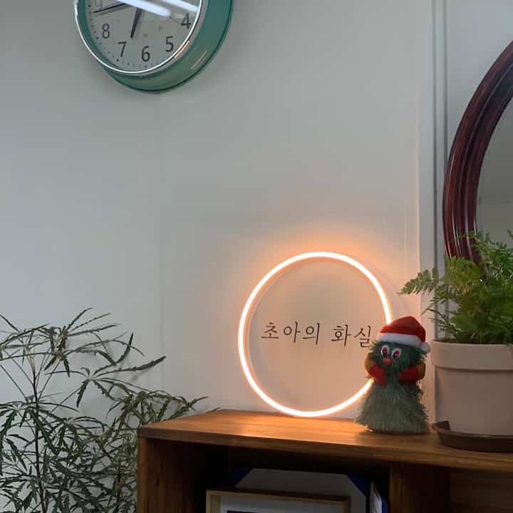 Natural wood tone shelf with circular neon sign, Christmas figurine, green rim clock on the wall, and plants in a cozy workspace