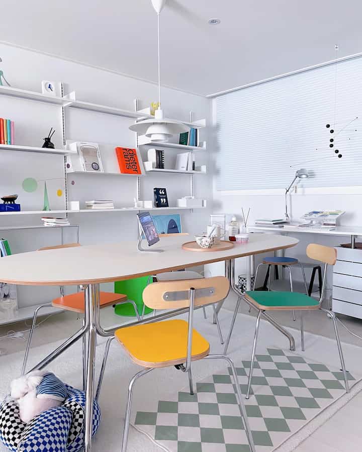 A white and pastel-toned living room featuring colorful chairs and a large desk with a modern atmosphere