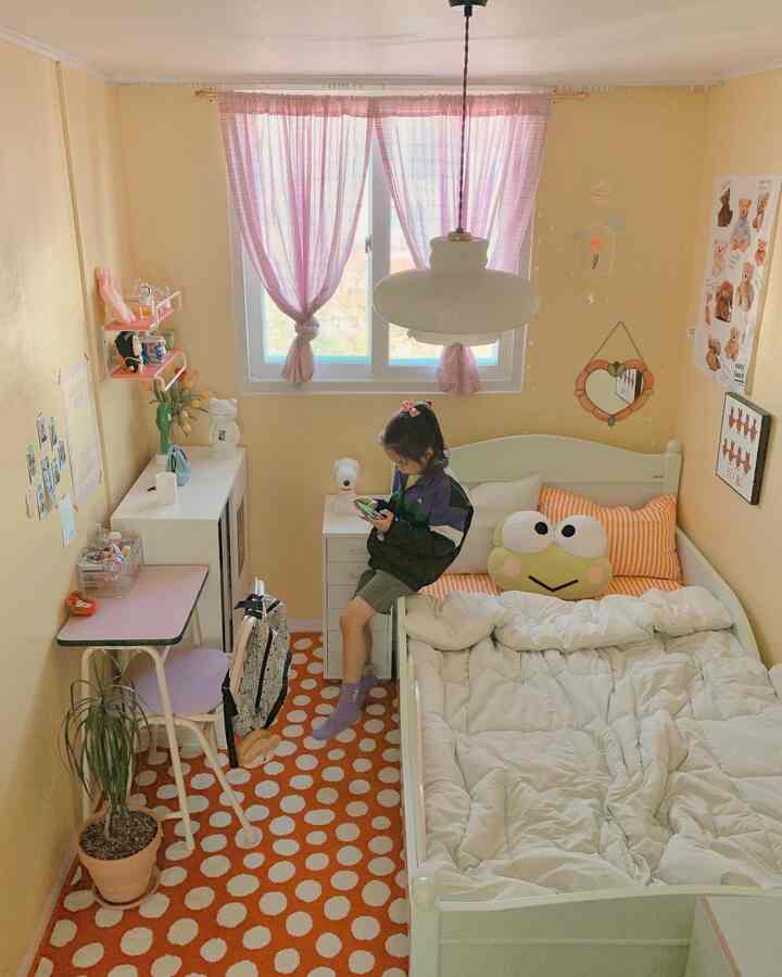 明るいイエローとホワイトの色調の子供部屋、白いベッドとオレンジのドット柄ラグがあり、子供がベッドのそばでスマホを使っている印象的な空間