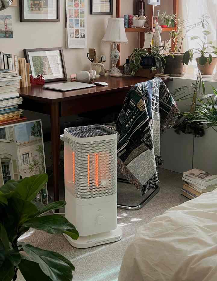 温かみのある色調のワンルームホームオフィス、白い電気ヒーターと木製デスク、植物が配置された居心地の良い空間