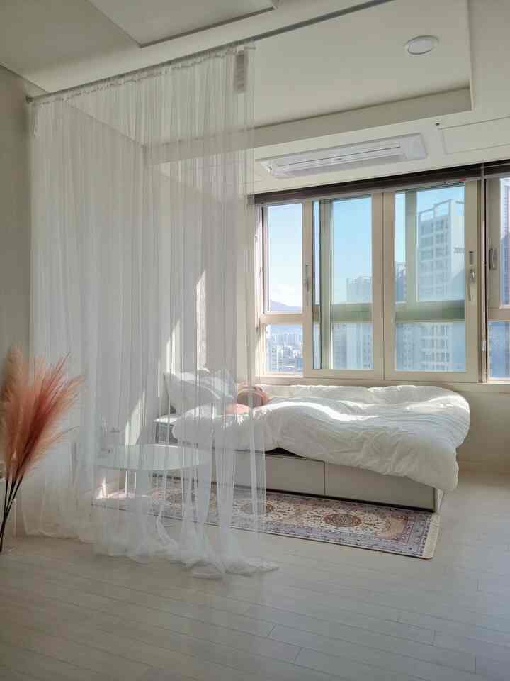 Bright white-toned studio bedroom featuring sheer mesh curtains and beige rug with natural decor accents