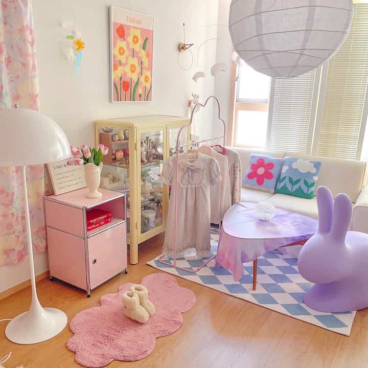 パステルピンクと紫のトーンが特徴のリビング、白いソファと花柄クッション、赤ちゃんの服が掛かるハンガーが温かみのある雰囲気を演出している