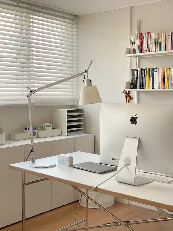 White and wood-toned home office space featuring clean desk and blinds, exhibiting a minimal and modern interior atmosphere