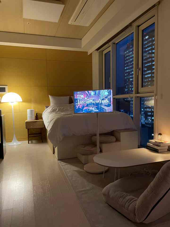 暖かい黄色い照明と木目調の壁が特徴のワンルーム寝室、ベッドとラタンのサイドテーブル、快適なアームチェアが配置された居心地の良い空間