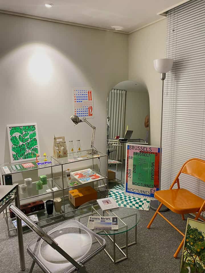 White and orange toned study featuring transparent chair, metal frame desk, and various posters with a modern aesthetic