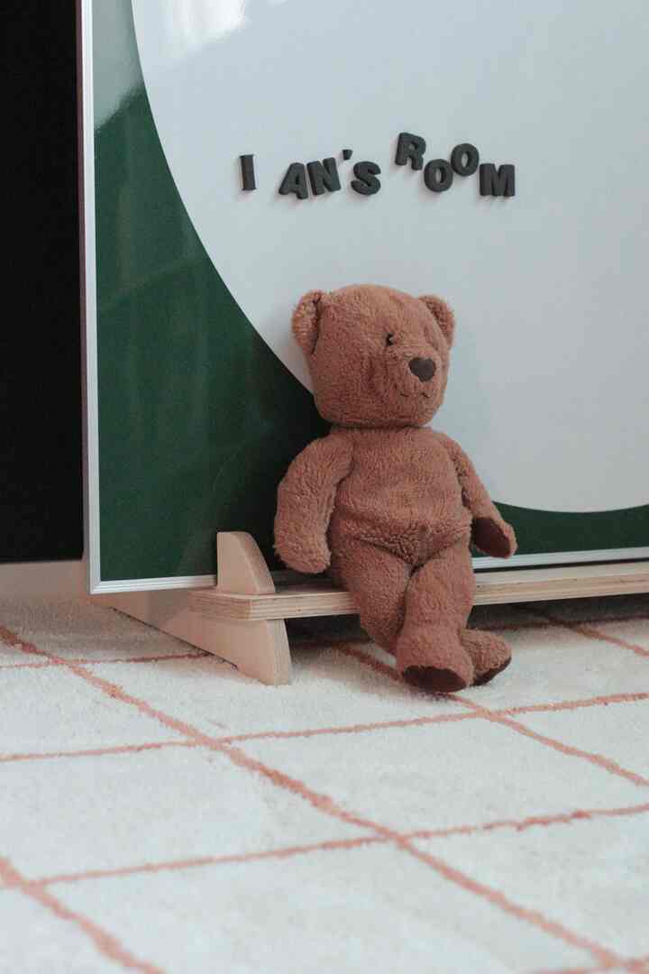 白と緑の色調の子供部屋、ピーチグリッドラグの上に茶色のテディベアが座る、落ち着いた雰囲気のあるインテリア