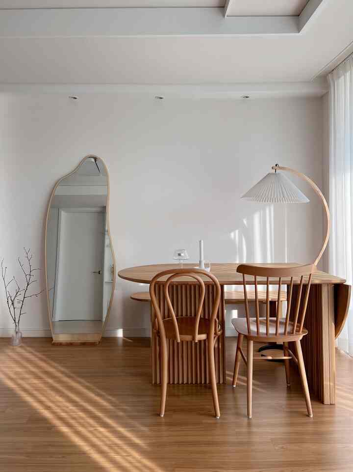 Wood tone natural living room featuring a wooden table, uniquely shaped full-length wooden mirror, and pleated floor lamp with simple ambiance