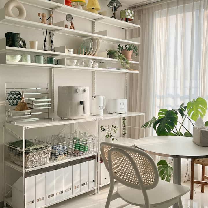 White and beige toned kitchen dining area featuring a round table, wall shelves with appliances and tableware in a cozy natural interior