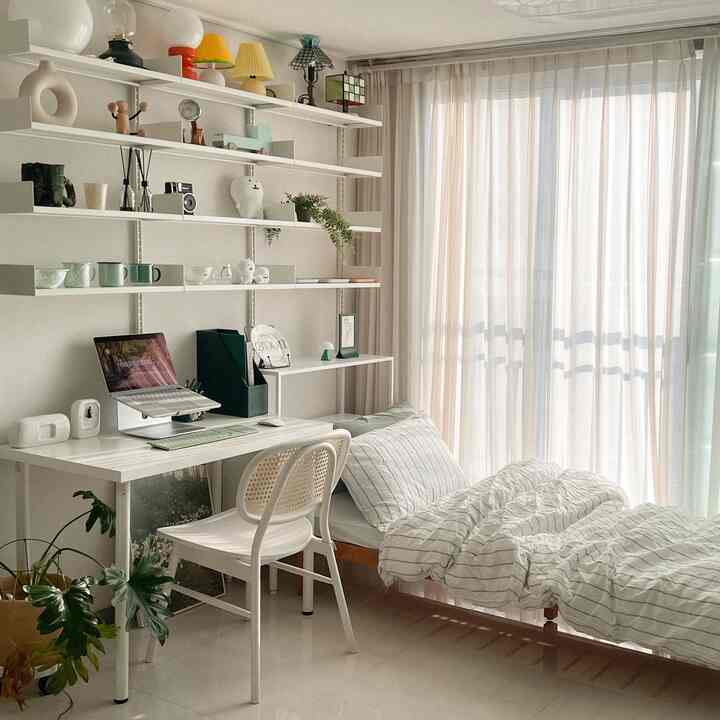 A cozy and clean single household bedroom in white and beige tones, featuring a desk and wall shelves