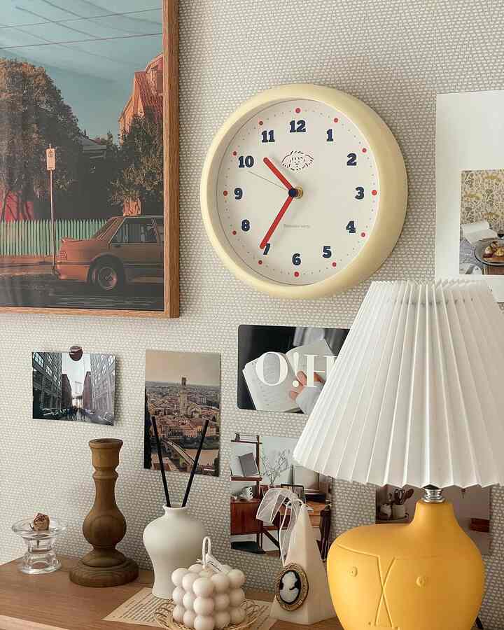 A cozy space featuring a beige and white toned desk with a yellow table lamp, various objets, a clock, and wall art