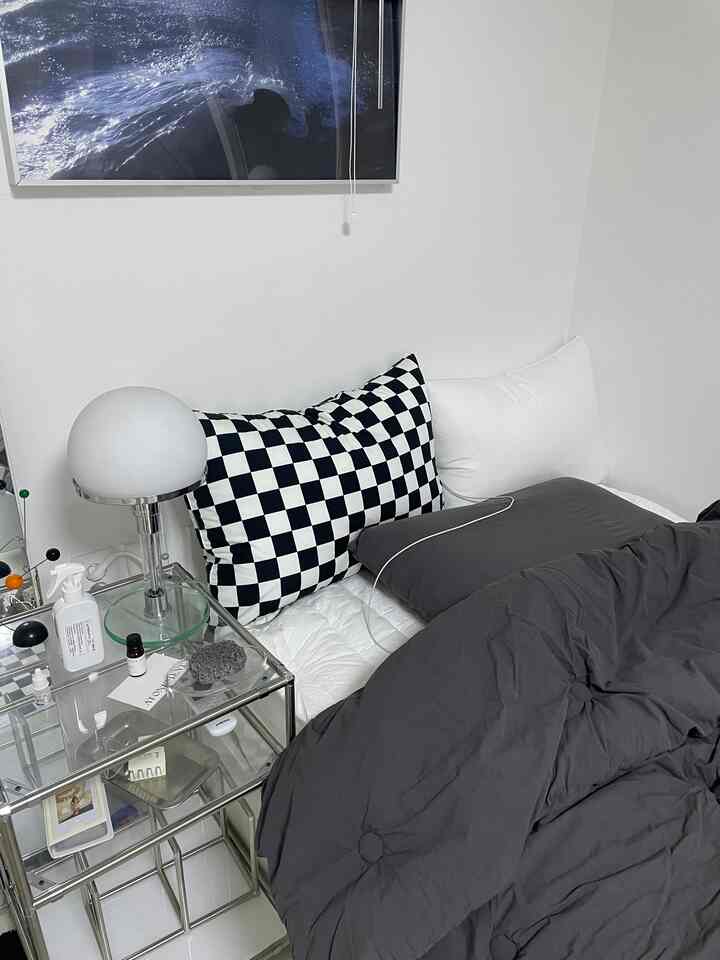 白い壁とグレーのベッドカバーが調和する寝室、モダンなメタルのナイトテーブルとチェッカーピローが配置された清潔感のある空間