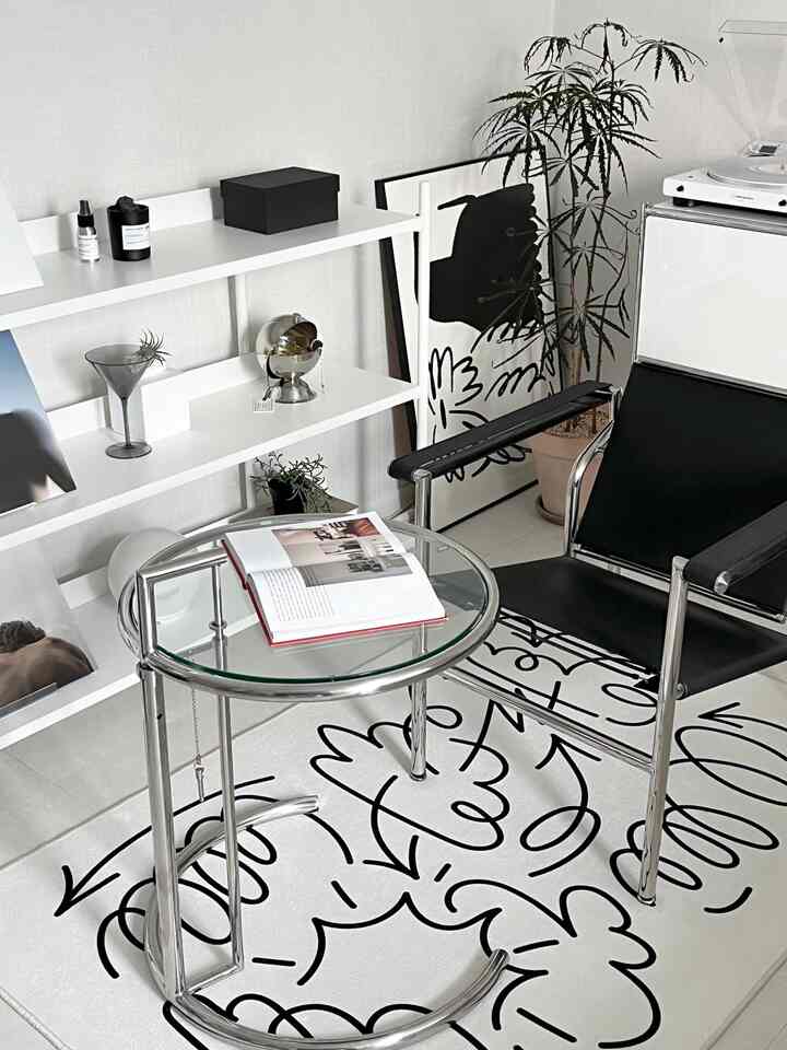 A white and black toned living room featuring a graffiti-style doodle rug, modern metal armchair, and glass side table in a clean minimalist setup