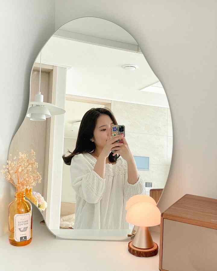 Bright and minimal vanity space featuring white and wood tones, an irregular shaped mirror, and an ambient table lamp creating an Instagrammable atmosphere