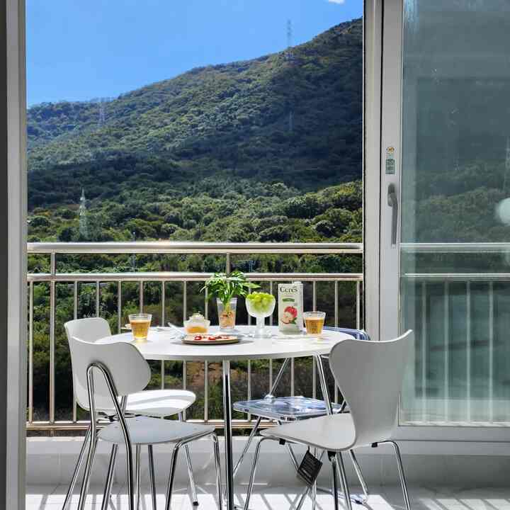 明るいホワイトトーンのバルコニー、丸いダイニングテーブルと透明な椅子が配置され、背後に緑豊かな山が広がる雰囲気のあるおうちカフェ空間