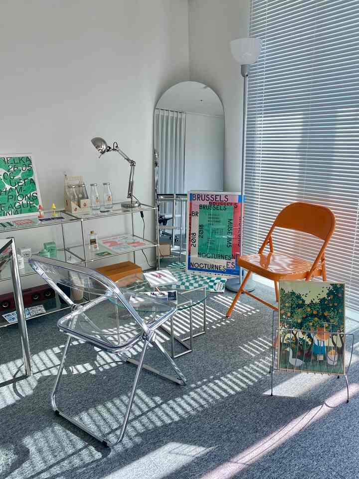 白とグレーのトーンのワンルーム書斎、透明な椅子とオレンジ色の折りたたみ椅子が配置されたすっきりとした読書・作業空間