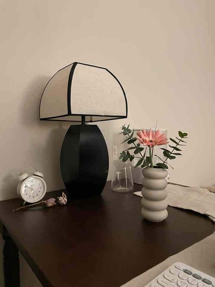 ベージュの壁とダークウッドのテーブルに、黒いテーブルランプ、白い目覚まし時計、ピンクの花が入ったベージュの花瓶が置かれた雰囲気のある空間