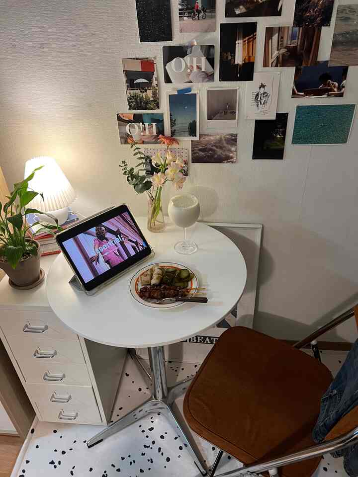 白い壁と丸テーブル、茶色の折りたたみ椅子と観葉植物、ポスターが配置された落ち着いた小さなリビング空間