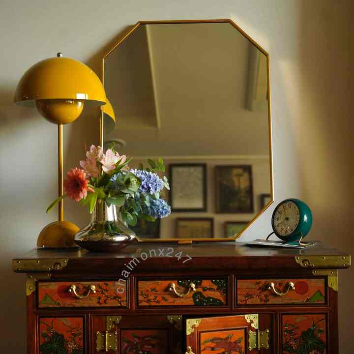 ゴールドトーンの八角形の鏡と黄色のテーブルランプ、カラフルな花瓶が置かれた伝統的な木製キャビネットの空間