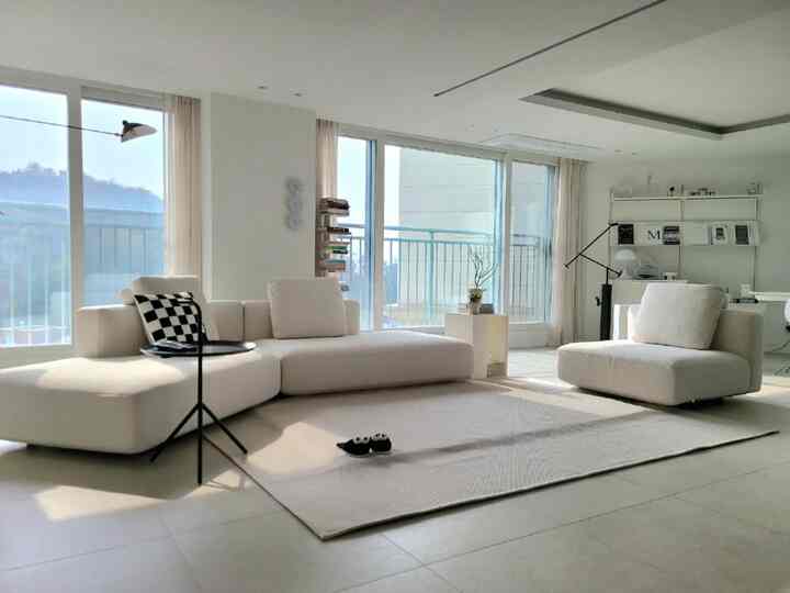 明るく清潔感のある白とアイボリー調のリビング、クリーム色のコーナーソファとラグ、広い窓が抜け感あるモダンなナチュラルインテリアを演出