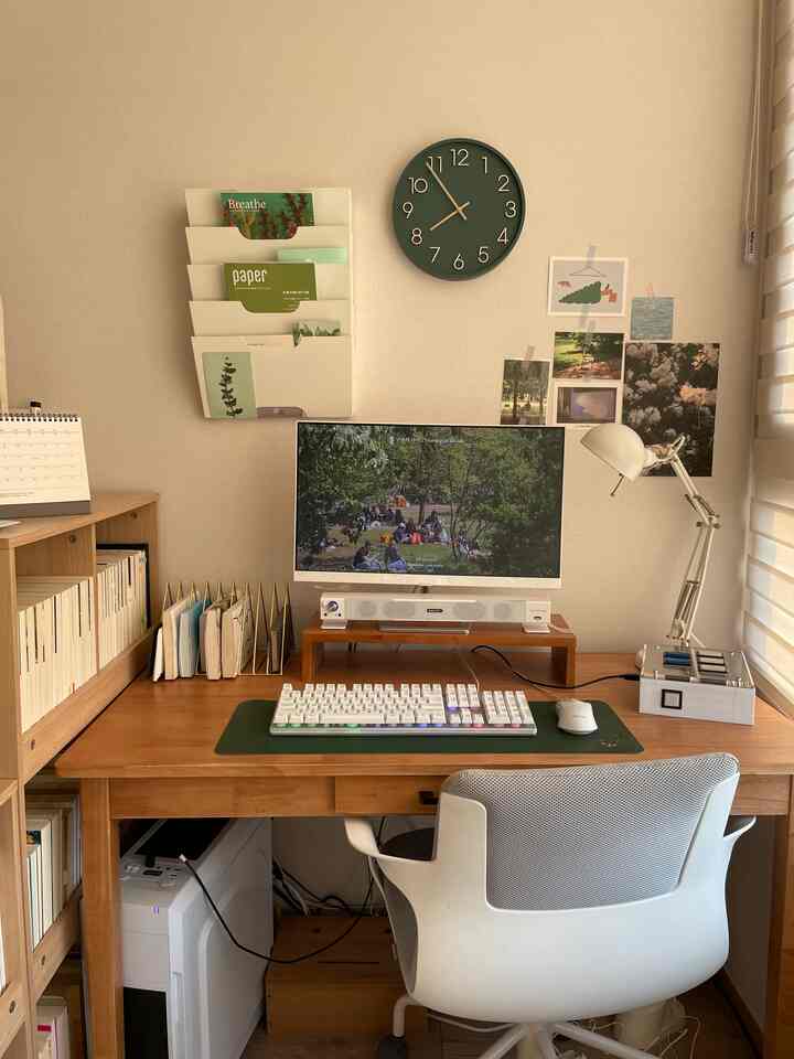 Natural wood tone home office featuring a central wooden desk and gray office chair, with a green clock and bookshelf on the wall.
